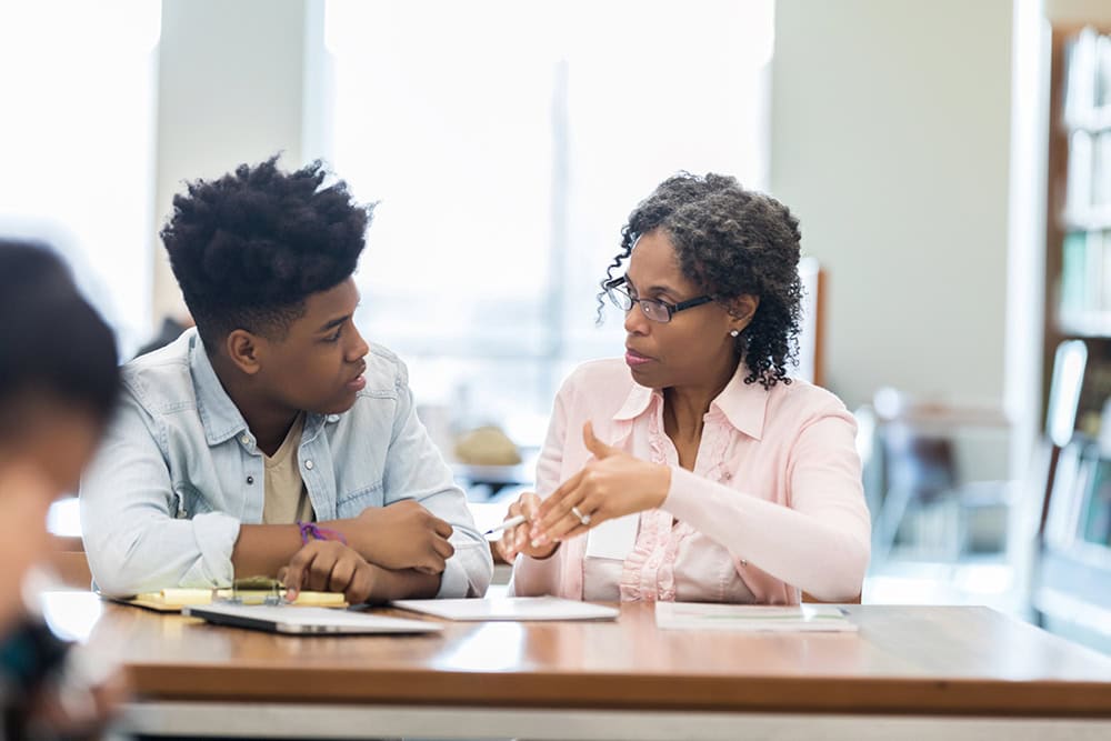 African American female volunteer tutor explains a math concept to a male high school student during a tutoring session.
