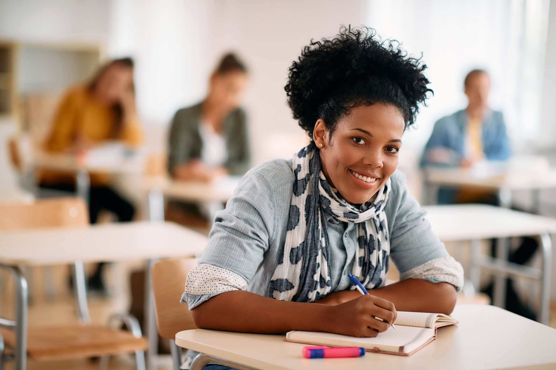 Happy Black woman writing an exam while attending a class at the university and looking at camera.