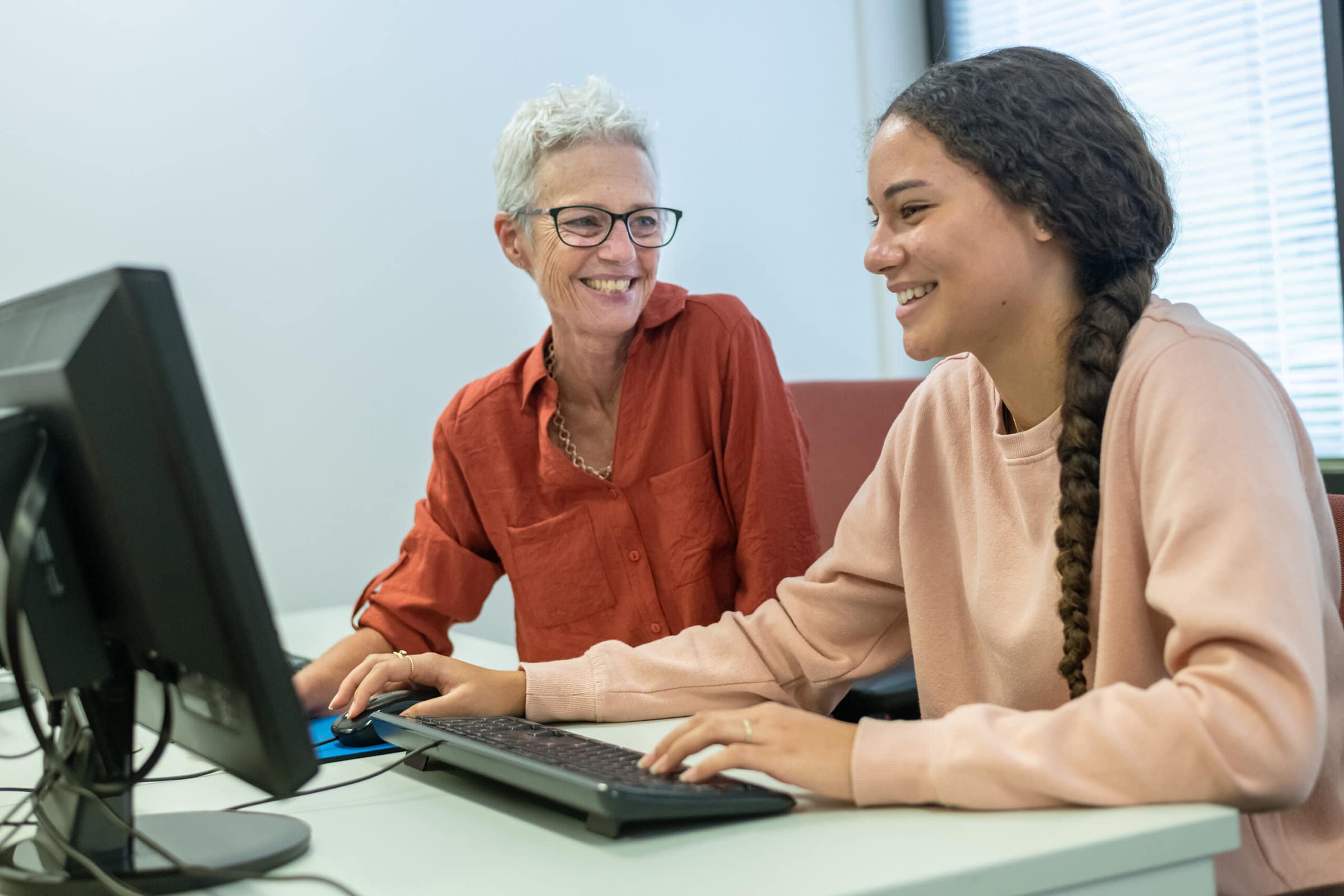 A teenage girl of Pacific Islander descent smiles while working on a computer in the school library with the help of a mature adult female teacher or tutor.