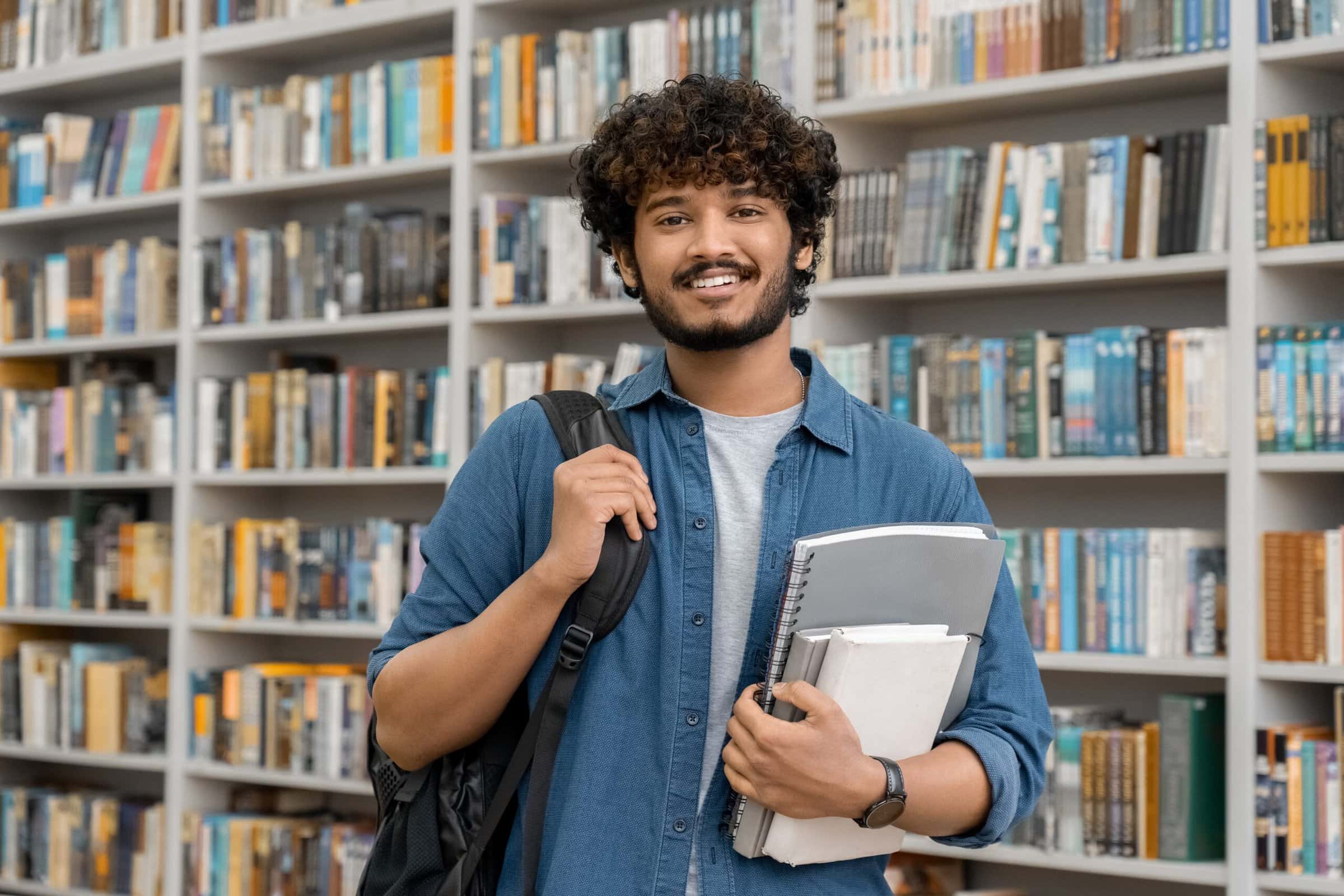 Portrait of cheerful male international Indian student with backpack, learning accessories standing near bookshelves at university library or book store during break between lessons