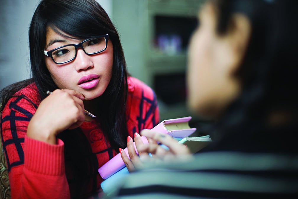 Indoor over shoulder view shot in study room of two late teen Asian girls talking to each other face to face. Selective focus is on the girl holding books wearing eyeglasses and listening carefully to her friend in foreground. Two people, waist up, horizontal composition with selective focus.