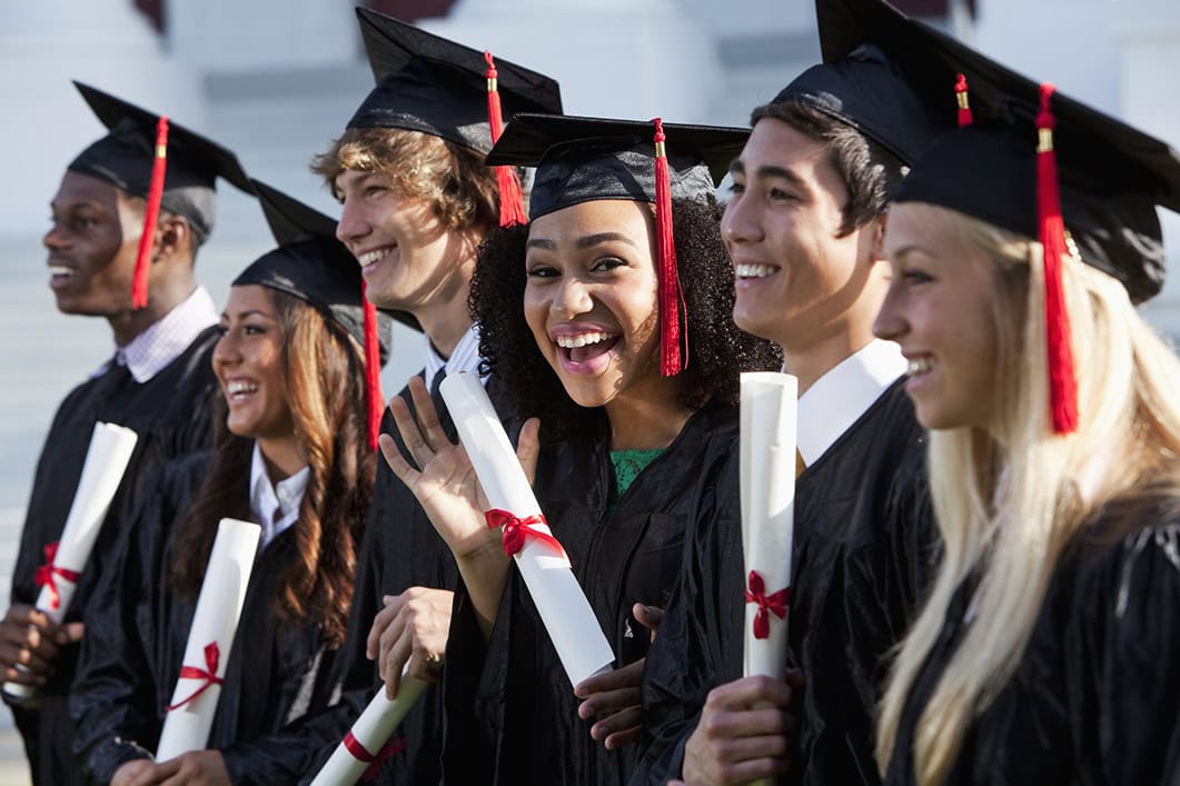 Multi-ethnic friends graduating together, in cap and gown. Main focus on African American girl in middle, waving at camera.