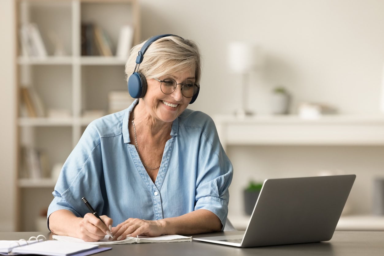 Happy senior woman in earphones and glasses studying on Internet from home, writing notes at laptop, watching online educating lecture, smiling, laughing
