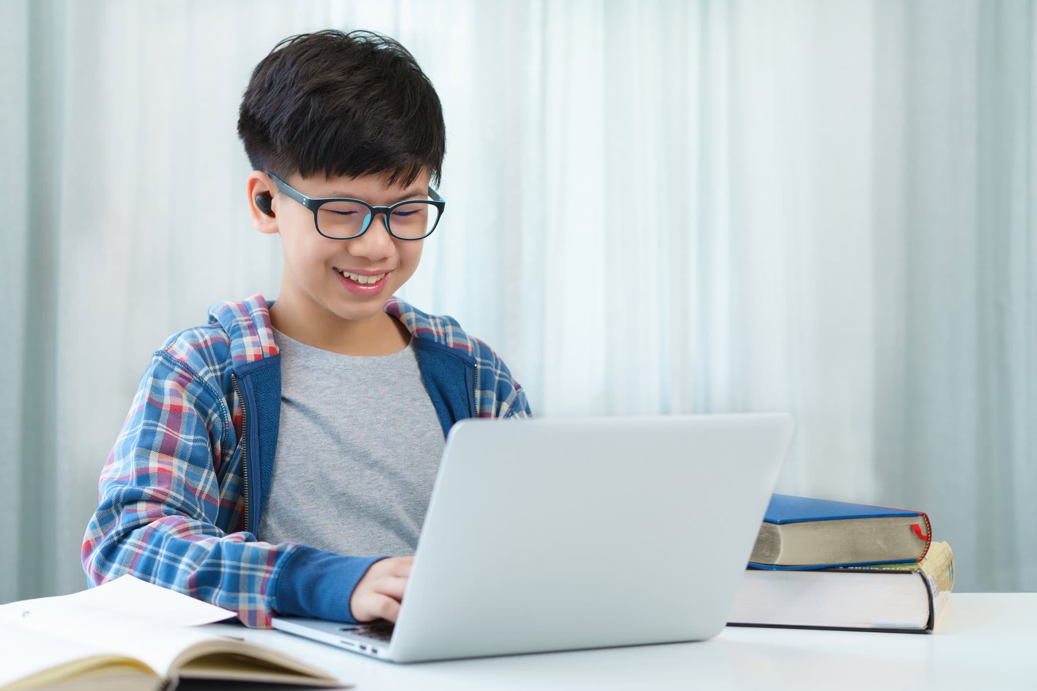 Happy Asian preteen boy with blue jacket and eyeglasses smiling, videocalling, learning online class with laptop computer in bedroom at home