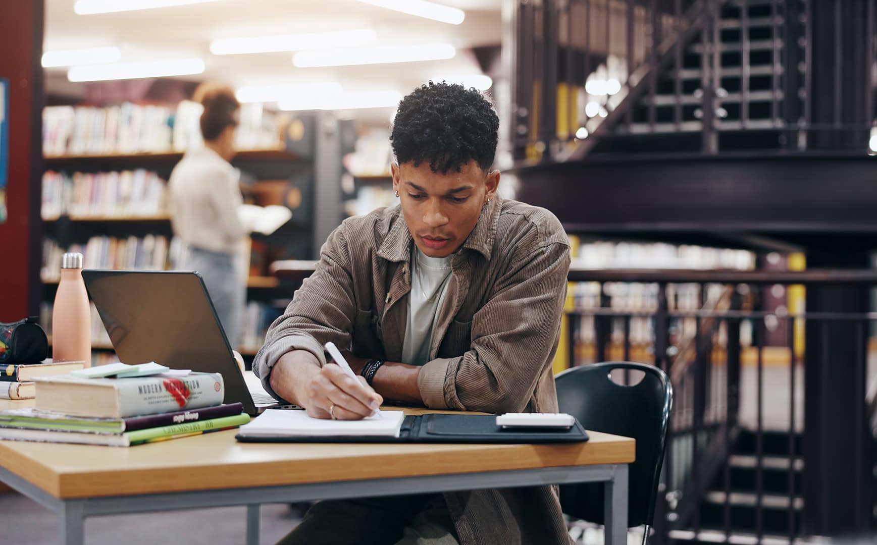 College student writing in library for research information, university thesis and study schedule. Male person, laptop and reading for education knowledge, history exam and project planning