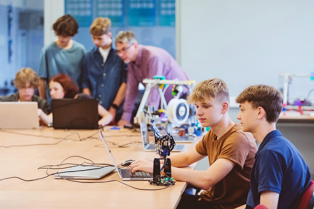 Gen Z teenage boys and girl high school students, coding movement of robotic hand at high school computer lab. Mature male teacher standing next to boys.
