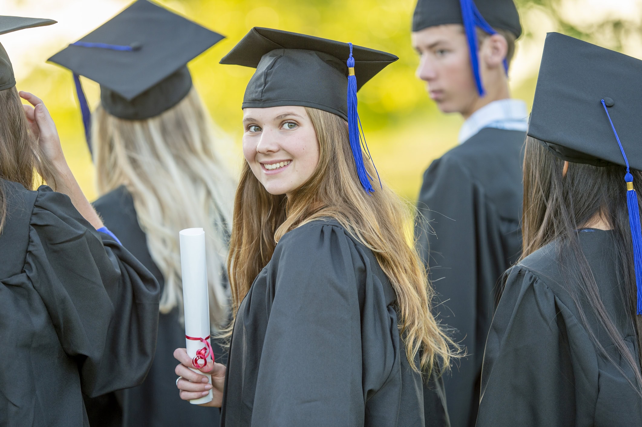A young female graduate turns around as she poses for a portrait. She is wearing a gown and cap and holding her diploma as she stands with her peers who have their backs to the camera.