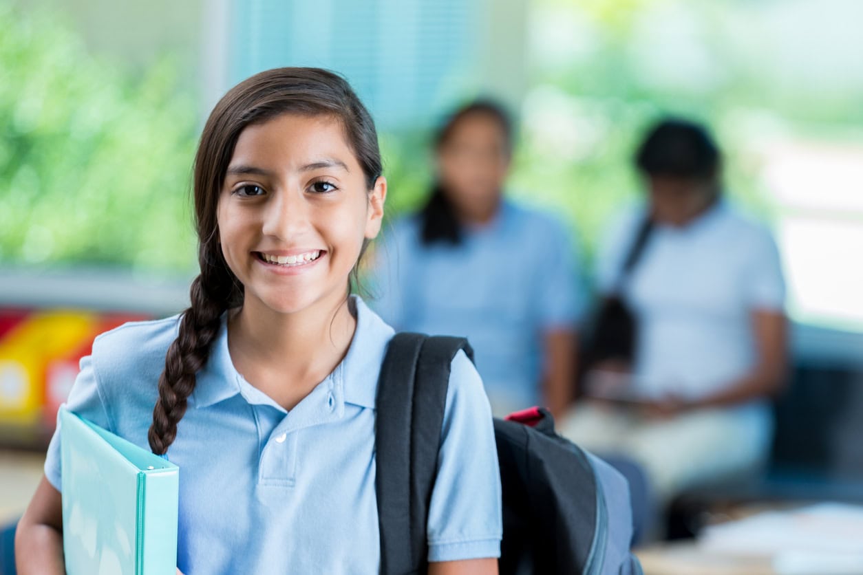 Cheerful private middle school student smiles before class. She is standing in her classroom. He has a braid and is wearing a backpack and a school uniform. Students are in the background talking.
