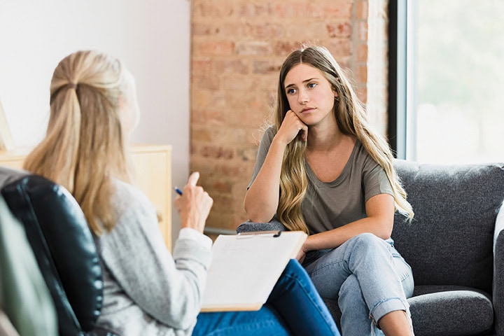 Teenage girl sits with her head resting on her hand as she listens to advice from the unrecognizable mature adult female counselor.