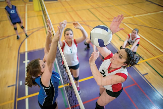 A group of teenage women playing in a volleyball match in a school gymnasium. One girl is jumping up to spike the ball.
