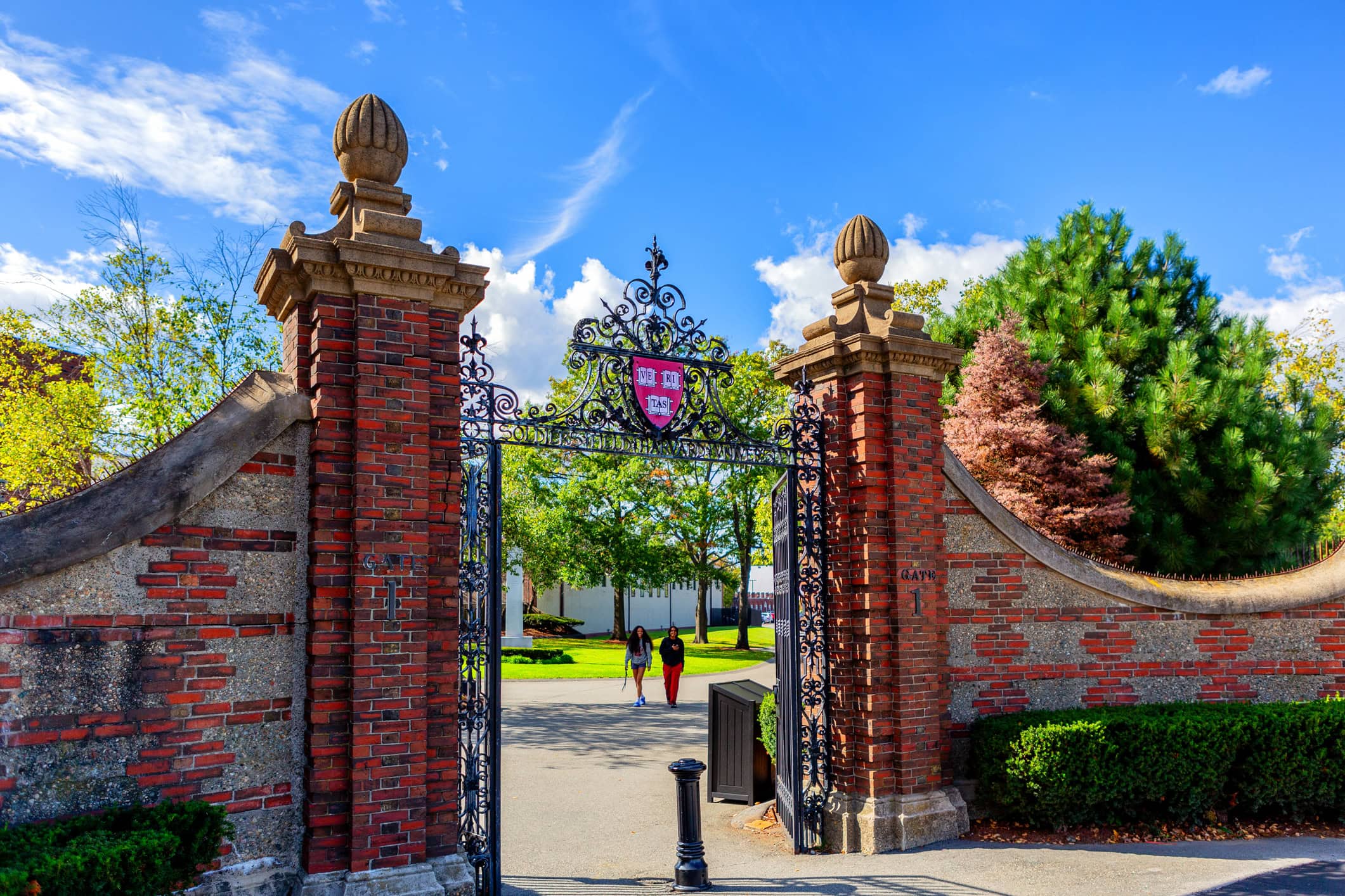 Boston, Massachusetts, USA - October 9, 2023: Gate 1, the Newell Gate pedestrian entrance to Harvard University's Soldiers Field Athletic Area. Atop the gate is the Harvard crest, logo, with three books and the word VERITAS (Truth). Two student pedestrians.
