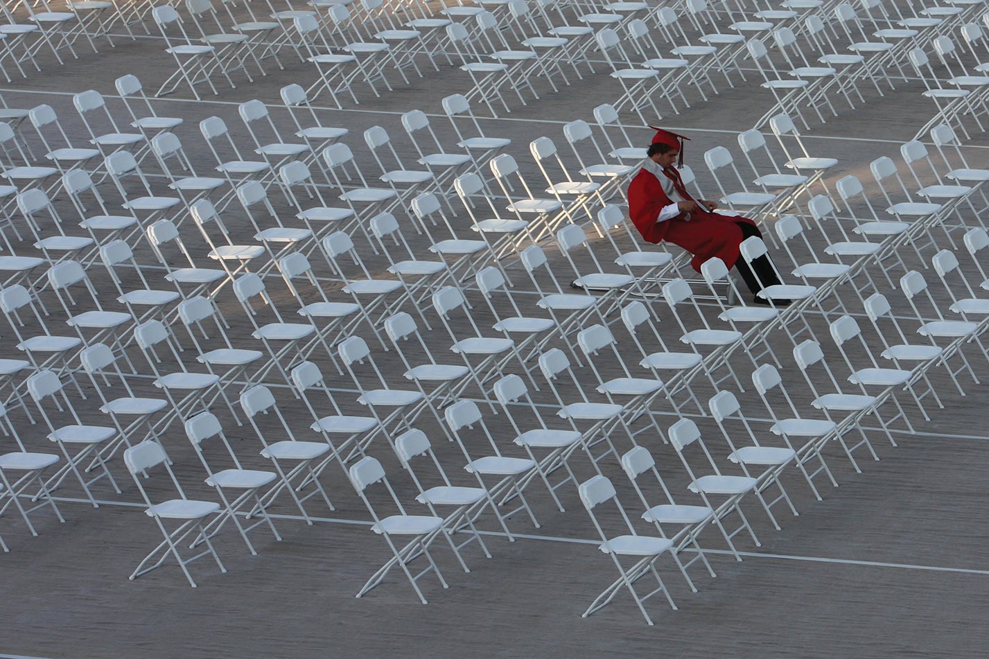A High School Graduate Sits Alone In Rows of White Chairs Waiting for Classmates