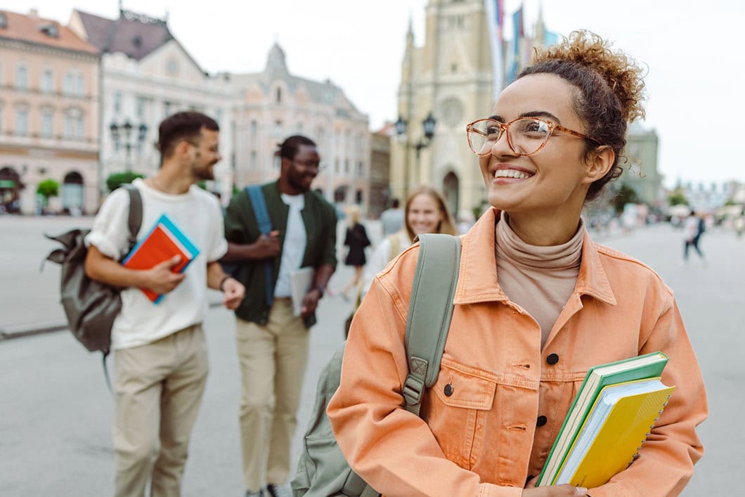 Young woman carrying books and standing in front of the University. A Group of other students is behind