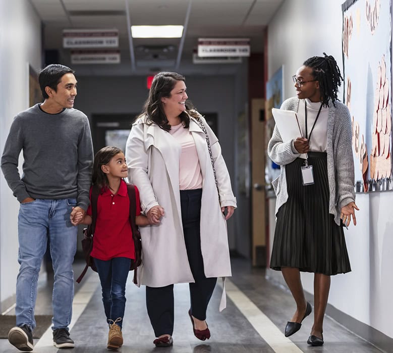 Two parents with their daughter on a school, talking with an elementary school teacher or principal in the hallway.