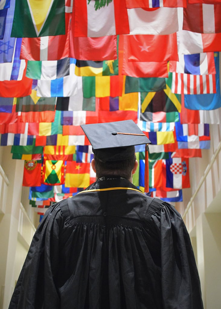 International student in grad cap and gown looking up at global world flags from different countries