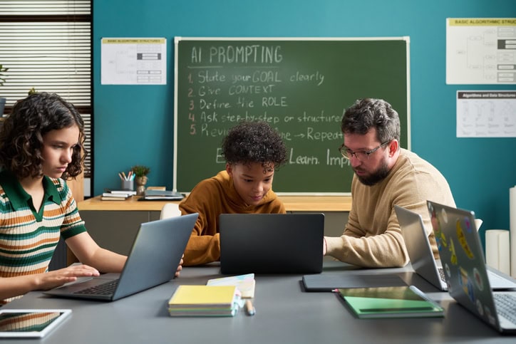 Teacher assisting students working laptops in classroom setting, chalkboard with AI prompting instructions visible in background