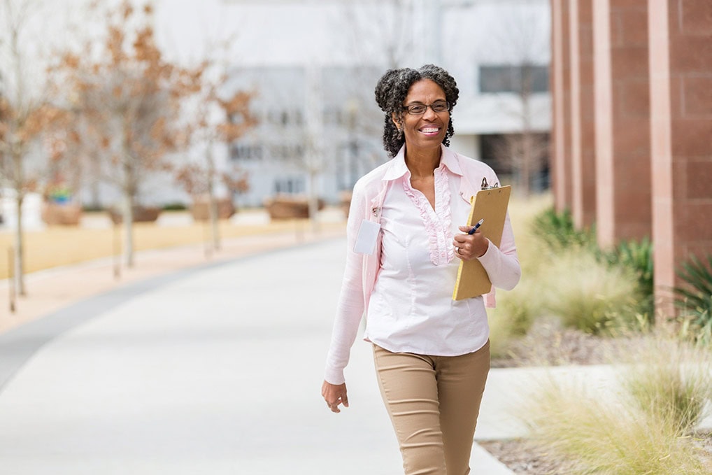 A cheerful mature female IEC walks along a sidewalk on campus. She looks ahead as she carries a clipboard and pen.