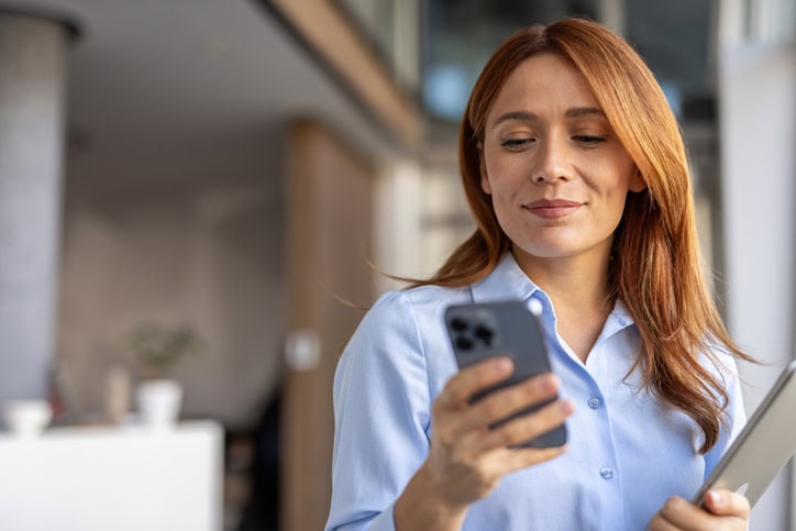 A business woman with a confident smile is standing in a modern office space looking at phone.